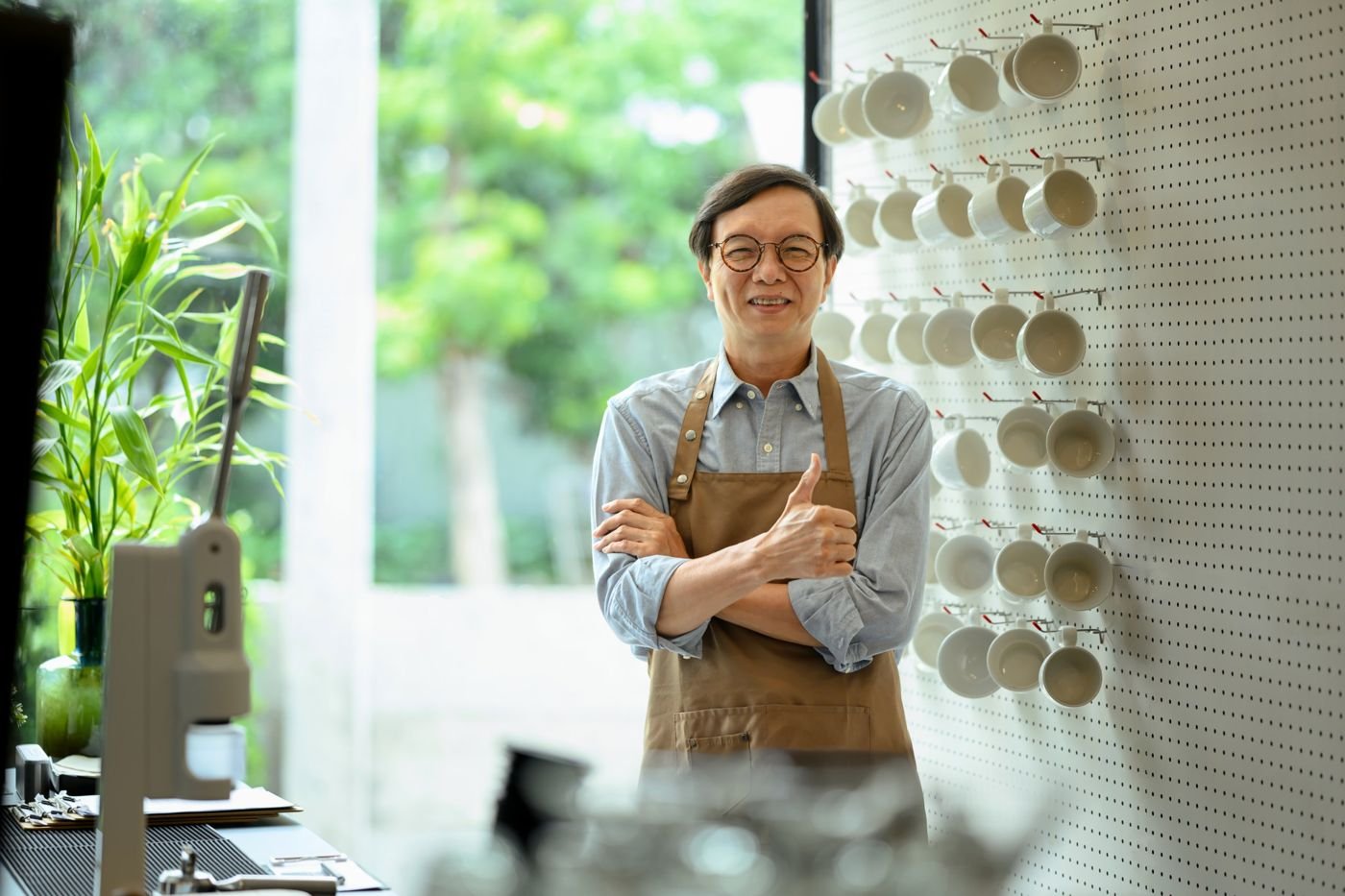 A smiling café owner in an apron giving a thumbs up beside a wall of hanging cups