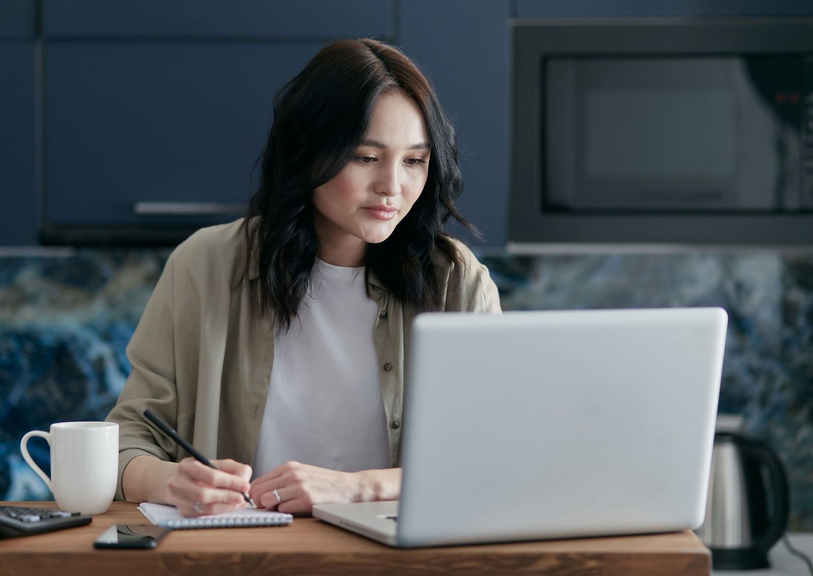 A woman making notes beside her laptop and a mug of tea at a wooden kitchen table