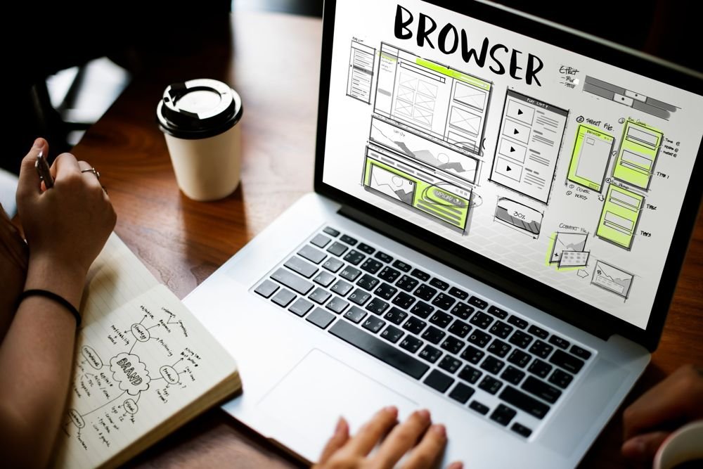 Hand-drawn website wireframes on a laptop screen beside a notebook of brand ideas and a coffee cup